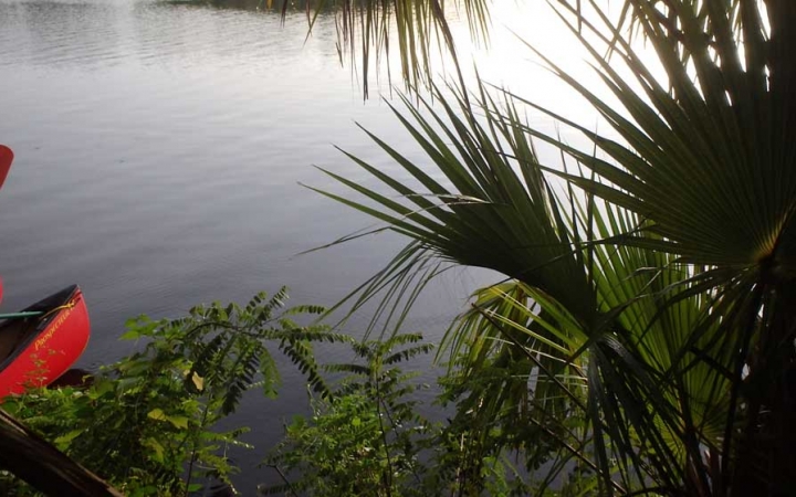 On the right side of the photo, tropical leaves frame a calm body of water. On the left side, the tips of two red canoes can be seen floating on the water. 